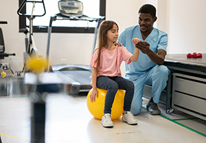 girl sitting on fitness ball while therapist holds her arm 